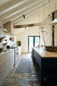 A beautifully renovated farm features a Sebastian Cox kitchen with a long cooker run, a large kitchen island with a double fluted Belfast sink, an aged brass Ionian tap, and a Carrara marble and solid oak worktop. Adorned with creamware pendant lights, vintage items, artisan ceramics, and a bowl of apples, this kitchen creates a clean and contemporary style that harmonises with the natural materials used throughout the space.