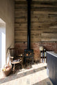 A quiet corner of a beautifully renovated farm features a Sebastian Cox kitchen and a cosy reading nook with a small burning wood cast iron fireplace, bookshelves, a vintage chair, and a basket of apples on a rustic original stone floor, along with exposed brickwork and wooden walls.