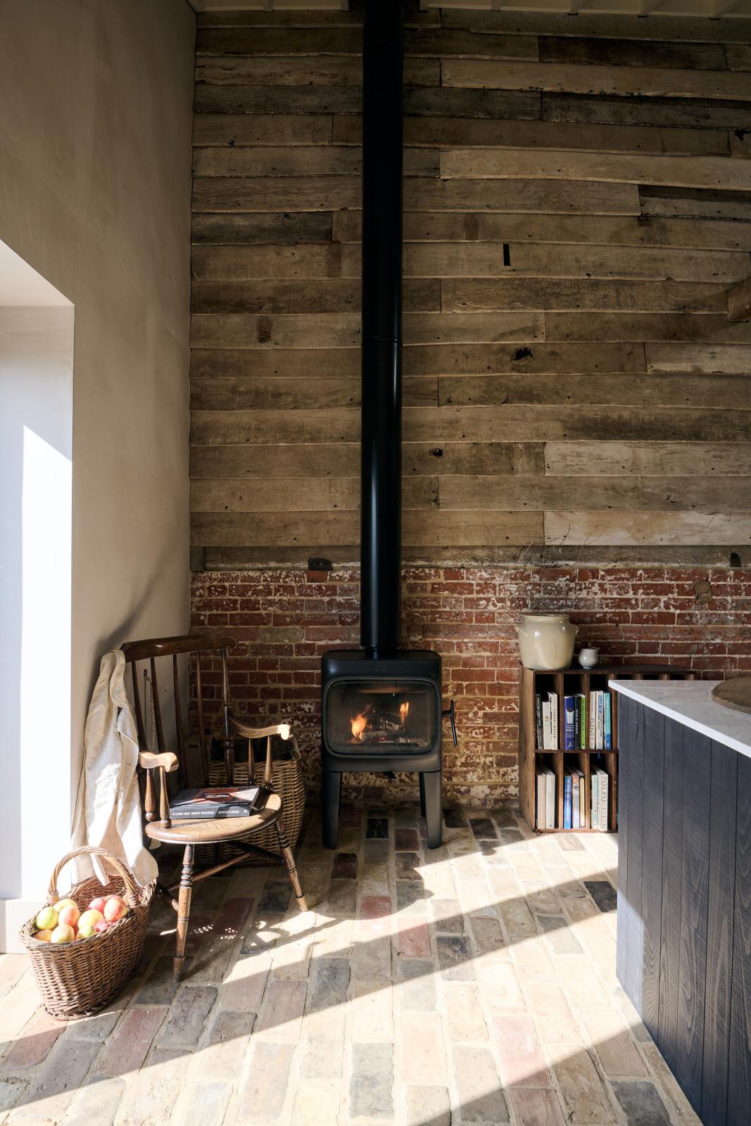 A quiet corner of a beautifully renovated farm features a Sebastian Cox kitchen and a cosy reading nook with a small burning wood cast iron fireplace, bookshelves, a vintage chair, and a basket of apples on a rustic original stone floor, along with exposed brickwork and wooden walls.