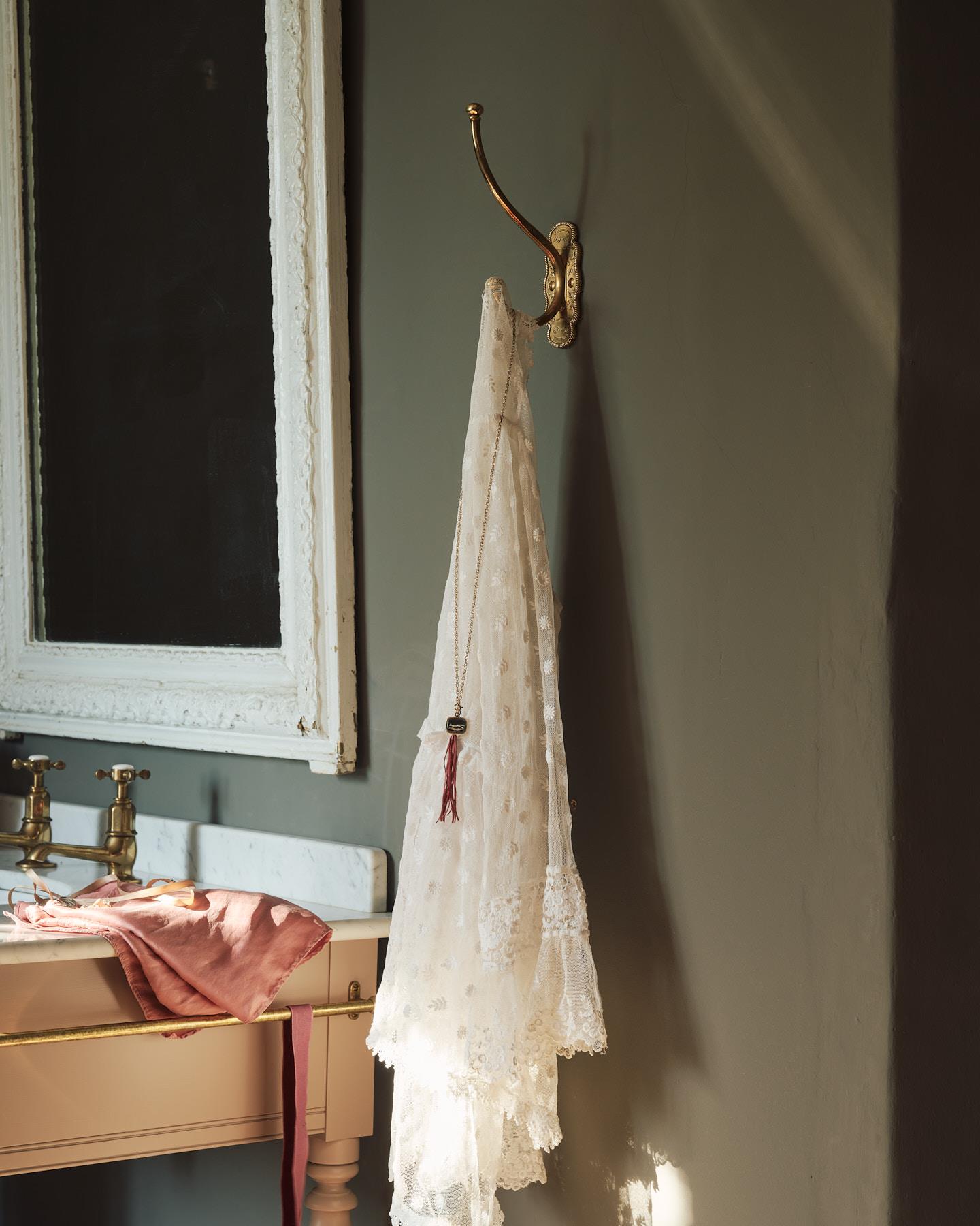A large Parisian café-style brass hook, fitted to a grey painted wall with a lovely vintage laced fabric and necklace draped on it.