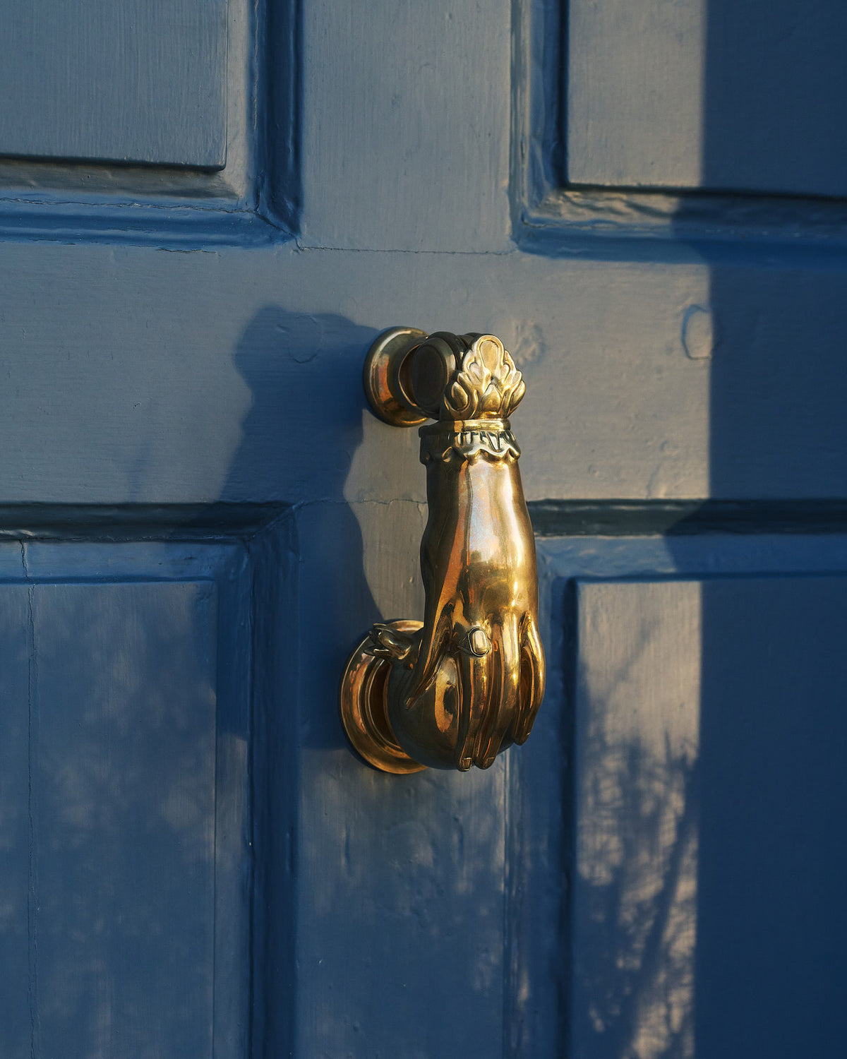 A decorative solid brass door knocker, shaped like a hand holding a pomegranate and fitted to a blue painted door.