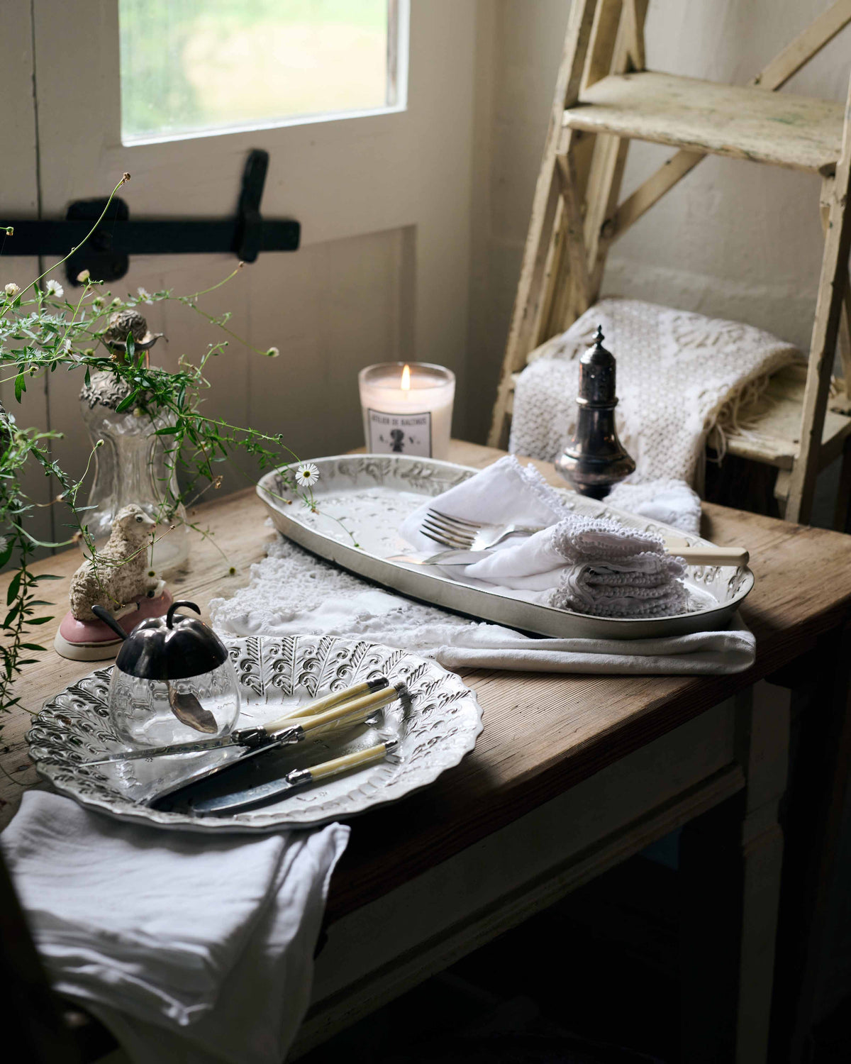 An oval and round handmade bohemian-style white ceramic platter with delicate impressions and surface patterns, complimented perfectly by vintage silverware, white linens and a rustic pine tabletop.
