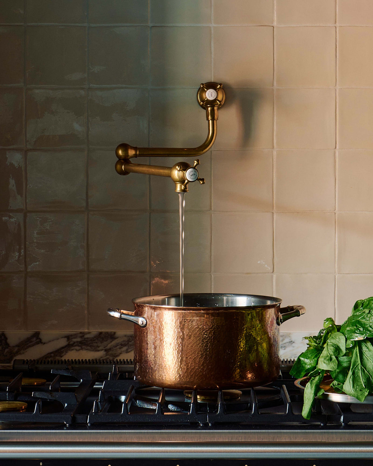 A close-up image of an aged brass pot filler tap attached to a cream-coloured splashback wall, filling a copper pot on a black gas stove with a marble worktop.