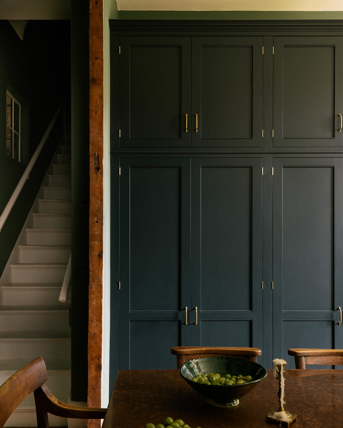 A bank of tall Georgian-style fitted pantry cupboards with a row of top cupboards above. The proportions and deep blue colour are classic and stylish, the simple cock bead on the door rails is highlighted perfectly by the soft sheen of the painted finish. A table with a mottled copper top, vintage wooden chairs, a bowl of gooseberries and a brass candlestick coated in melted wax sits in the foreground, the ruddy tones are a satisfying contrast to the cabinets.