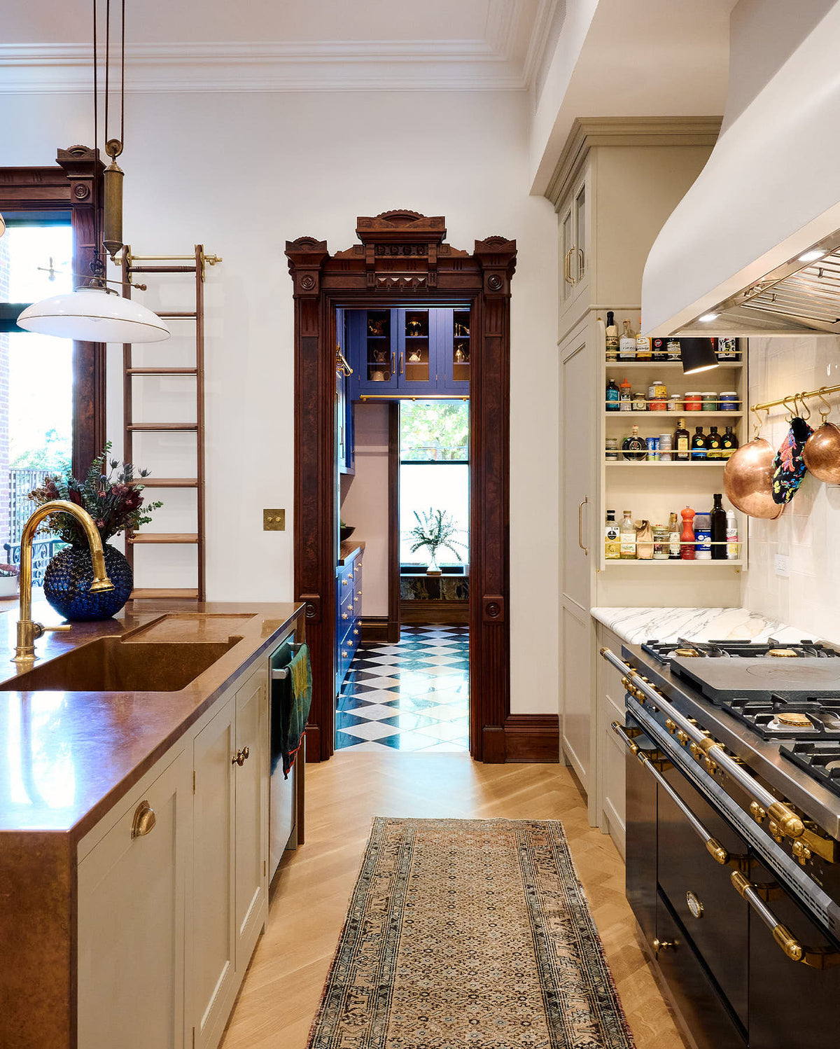 Past a grand and spacious classic English kitchen features a half vintage and half deVOL kitchen island with a copper sink and a cooker run with an Arabescato marble worktop is a blue walk-in pantry. Beautifully decorated with artisan glass and ceramics, a sophisticated floral arrangement, an aged brass hanging rail, a bespoke ladder, and milk glass pendant lights, it creates a social space with a touch of luxury.