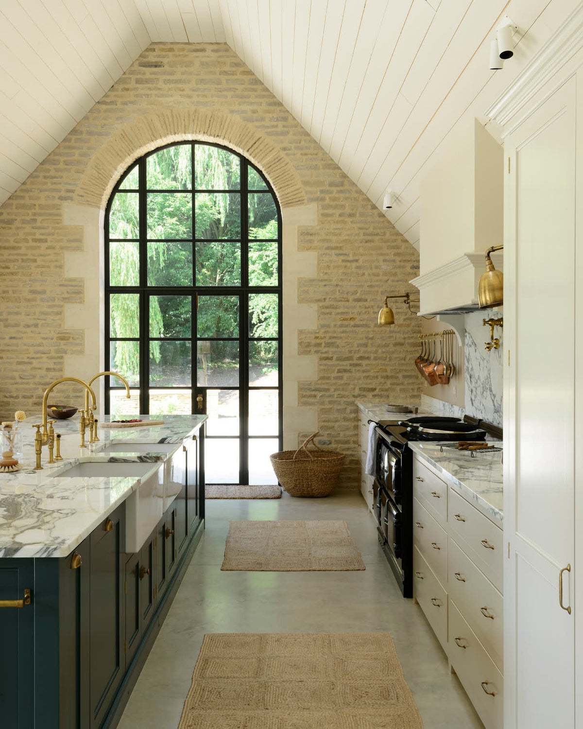This grand mushroom-coloured classic English kitchen features a cooker run with a suspended chimney breast, a large kitchen island topped with Arabescato marble, and a bank of full-height cupboards. Beautifully decorated with woven rugs, a vintage basket, simple ceramics, brass folded wall lamps, and a large collection of kitchen utensils, this space creates a humble family atmosphere despite its luxurious design.