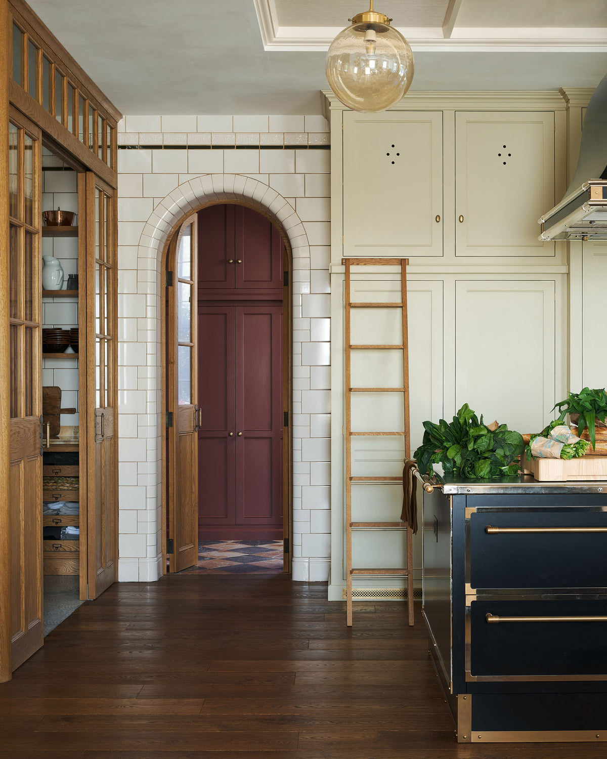 One side of a grand room—with a high panelled ceiling, glossy white tiled walls, and curved archways flanking a huge bank of creamy mushroom full-height classic cupboards with a polished oak ladder—features a black kitchen island with gold accents and a canopy. Beyond, an arched doorway leads to a refectory-red room housing the large haberdasher’s pantry.