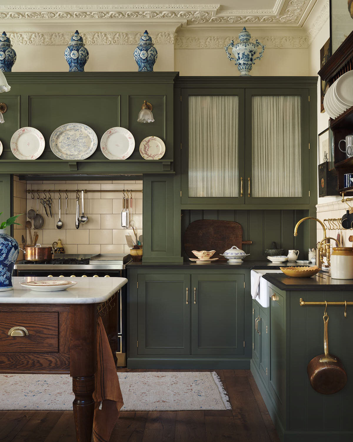 A quiet corner of an eccentric yet grand Bakehouse Green Classic English Kitchen features an L-shaped sink and cooker run with black granite worktop, a glazed wall cupboard, an overmantle with cream subway splashback tiles, and an Heirloom dairy table. Pridefully displaying taxidermy, artisan ceramics, and collectable antiques, it embraces a unique museum-like social kitchen space.