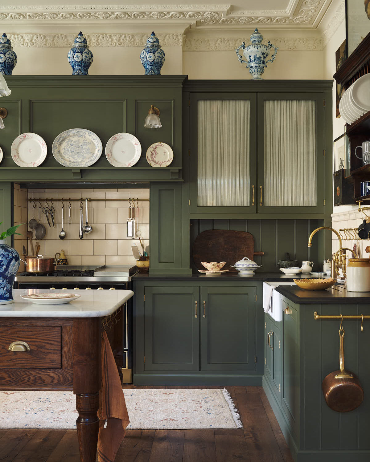 A quiet corner of an eccentric yet grand Bakehouse Green Classic English Kitchen features an L-shaped sink and cooker run with black granite worktop, a glazed wall cupboard, an overmantle with cream subway splashback tiles, and an Heirloom dairy table. Pridefully displaying taxidermy, artisan ceramics, and collectable antiques, it embraces a unique museum-like social kitchen space.