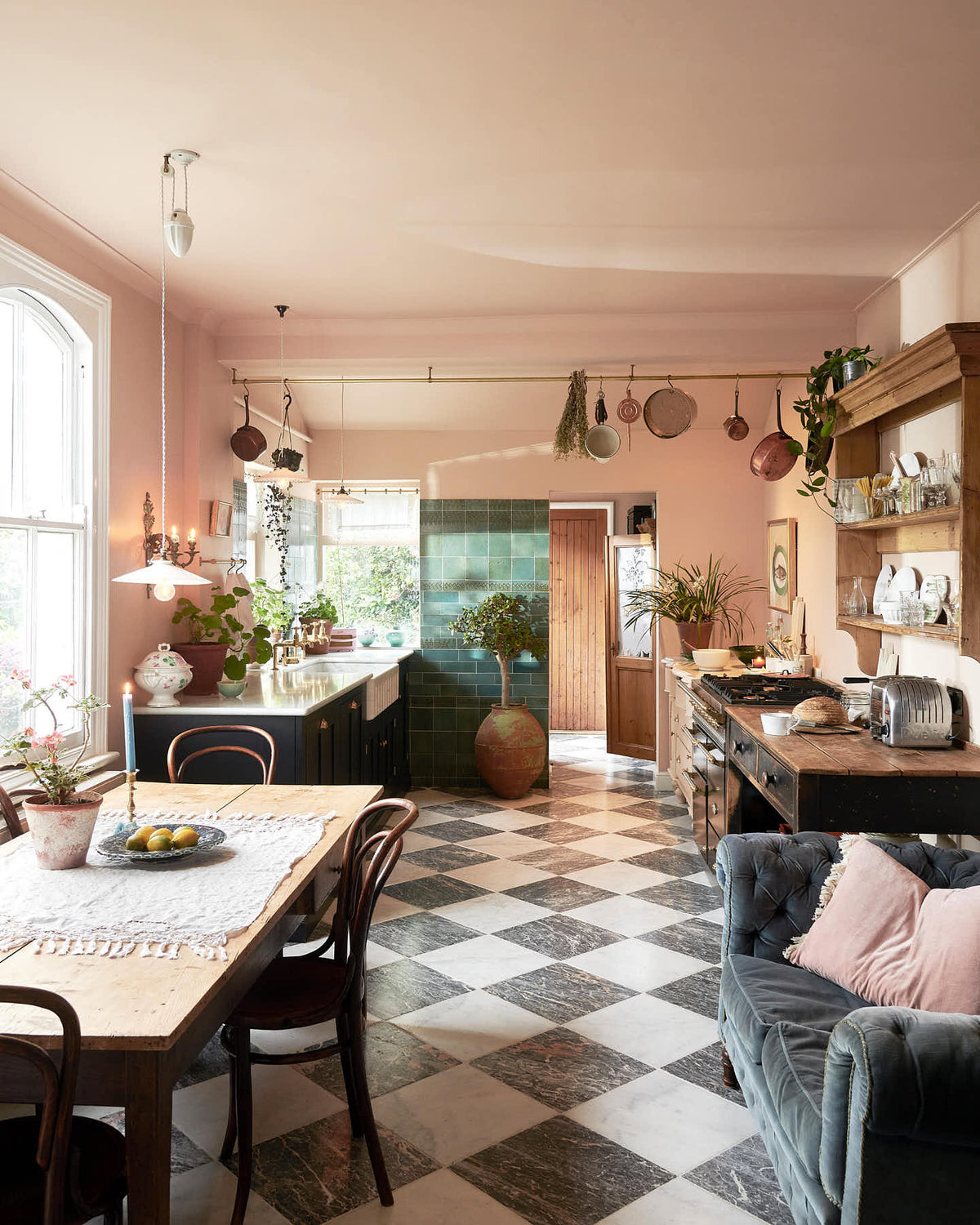 A bright peach-coloured room with a chequerboard marble floor and an eclectic collection of vintage furniture, including a table and chairs, a prep table and a decorative set of drawers. In the left corner is a modest sink run with black painted Classic English cupboards, a Carrara marble worktop and brass fittings. Pots and pans hang from a brass ceiling rail, mixed with a collection of vintage milk glass pendant lights.