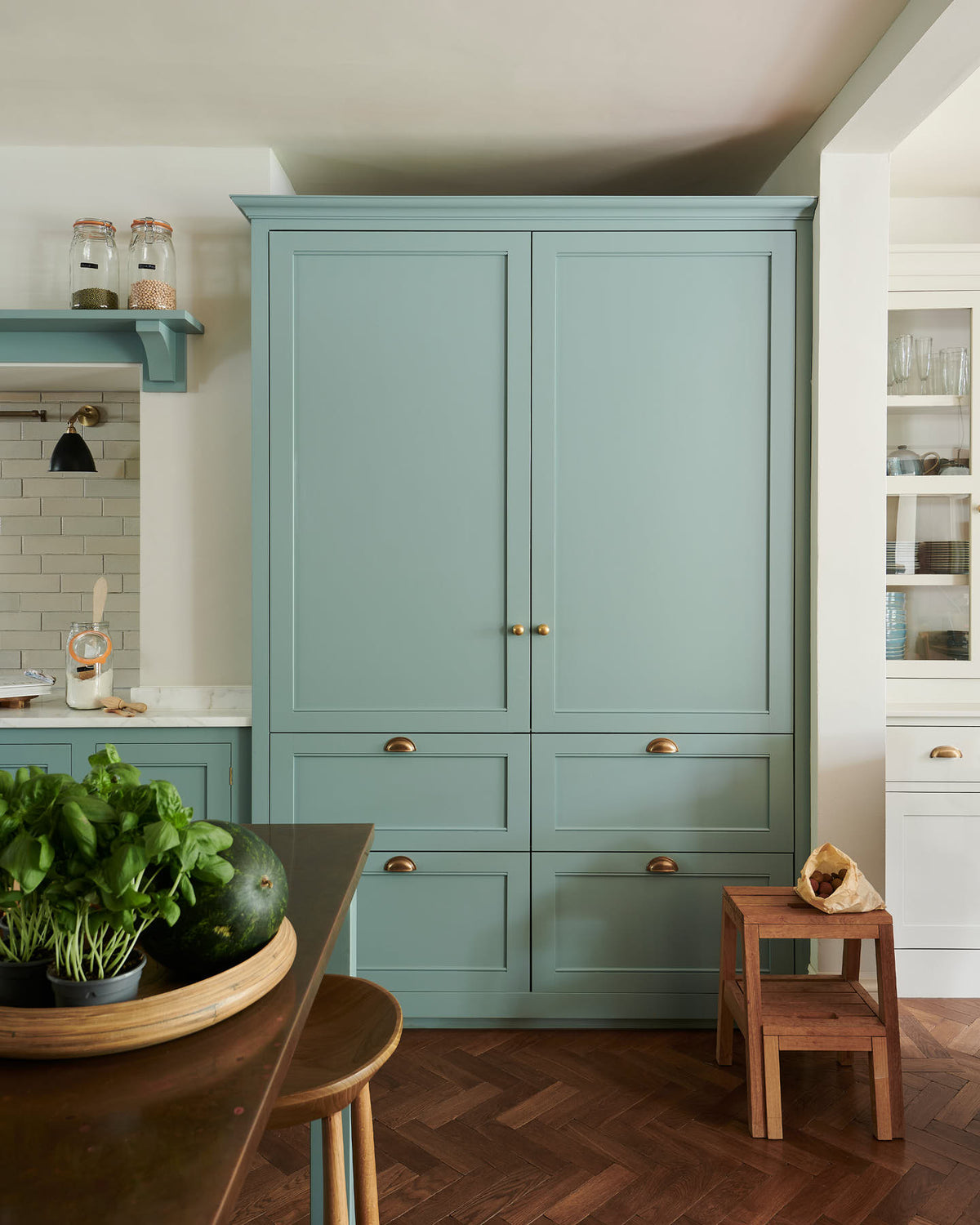 A spacious yet modest Trinity Blue classic English kitchen features a silestone worktop cooker run, an over mantle canopy with a white porcelain splash back, a large full-height cabinet housing a fridge/freezer, a copper prep table, and deVOL wooden bum stools. Lovingly decorated with a watermelon, live basil, and dried ingredients in glass jars, the room feels humble, warm, and inviting.