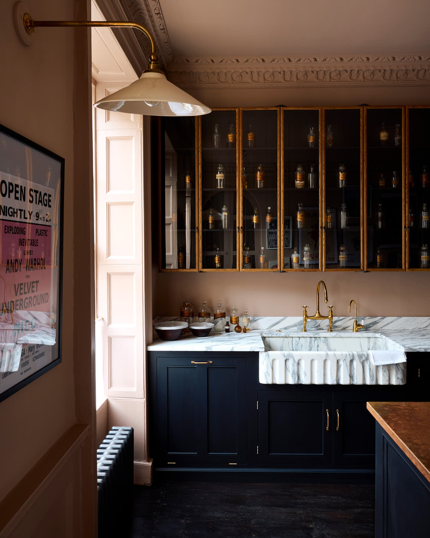 A quiet corner of a bold and dramatic Haberdasher’s kitchen with a sink and cooker run, an Arabescato worktop and single fluted sink, an aged brass Ionian tap, a glazed wall cupboard, and a kitchen island with a copper worktop. Adorned with a milk glass task light, artisan ceramics, labelled glass jars, and a framed poster, creating a local bristo bar in Manhattan space.