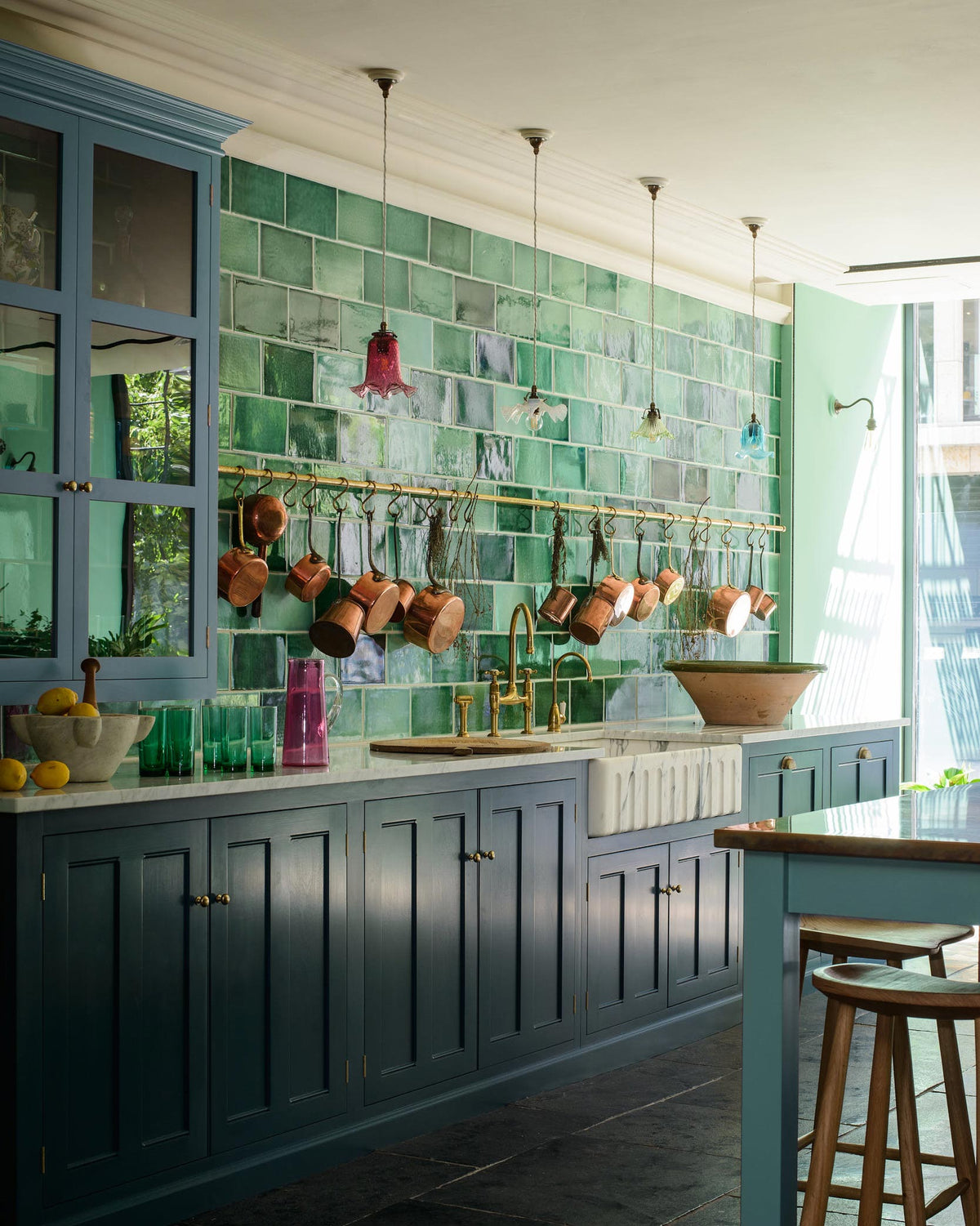 An angled view of a stunning, Bond Street blue kitchen features a sink run with Arabescato marble worktop, complemented by an Arabescato marble fluted sink fitted with an aged brass Ionian tap. The room features a stunning mix of colours and character, accentuated by colourful glass pendant lights, aged brass rails with copper pans, and handmade reclaimed green porcelain tiles from England.