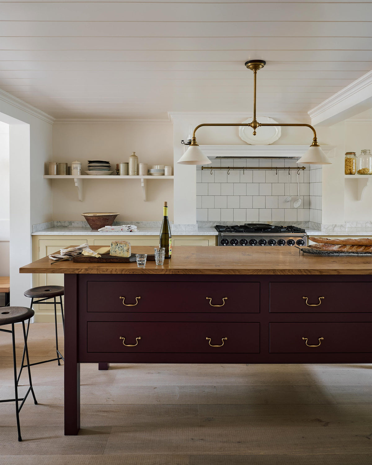 A warm, inviting, muted yellow classic English kitchen features an L-shaped cooker and sink with a Carrara marble worktop, a large freestanding maroon prep table with a solid oak worktop and a breakfast nook. Adorned with deVOL dark creedy stools, cherished muted ceramic plates, bowls, a deVOL brass heirloom gaselier light, freshly baked baguettes, wine, and a charcuterie board, this kitchen feels humble and inviting for a family that enjoys cooking.