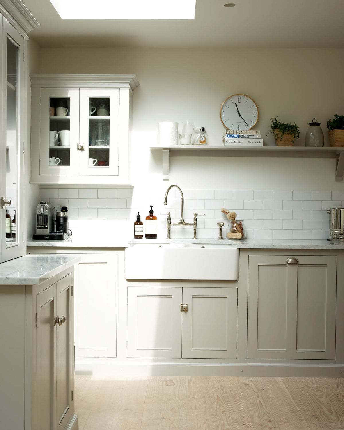 A close-up of a simple and stylish classic English kitchen’s double Belfast sink with antique silver Ionian tap, Carrara marble worktop, glazed wall cupboard, and shelf. Beautifully decorated with light copper rimmed clock, glass containers, wooden cleaning equipments, and white splash back tiles, it creates a functional and beautiful kitchen space.