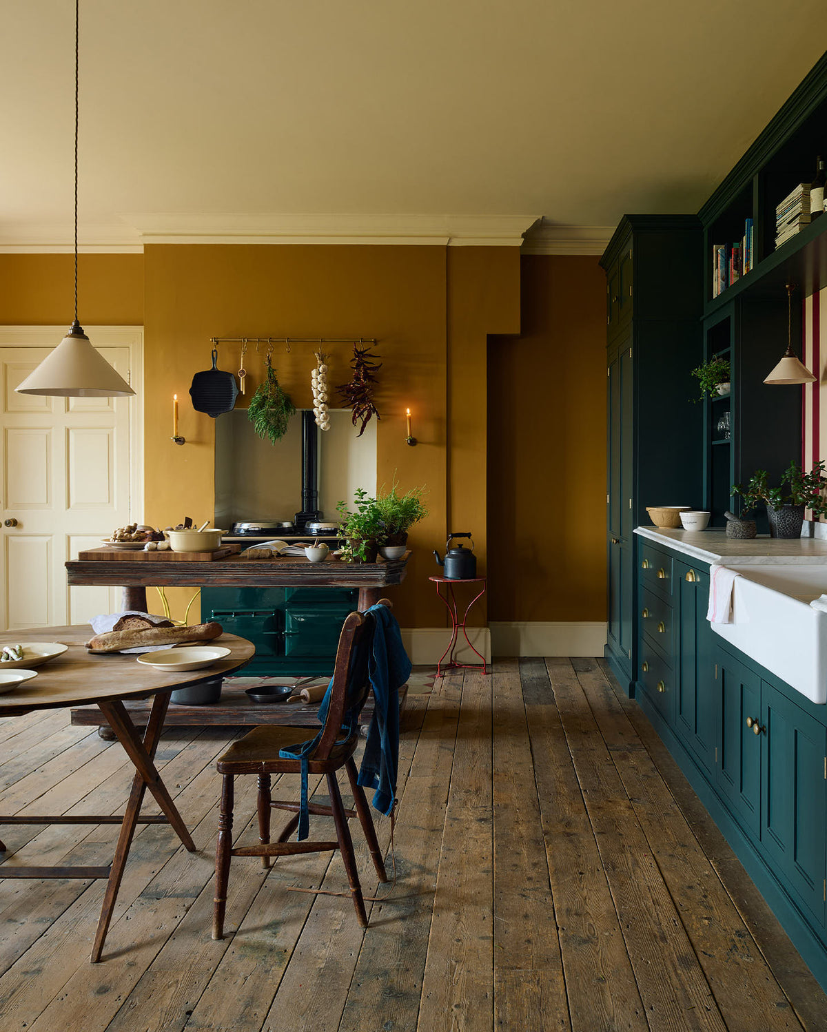 A lovely modern kitchen that retains the charm of its original structure and flooring, using the three primary colours in a muted, mature palette. The spacious kitchen features a Clerkenwell Blue full-height pantry cabinet, a long sink run with a marble worktop, and an open countertop cabinet and shelf. An emerald green stove sits within the chimney breast, accompanied by a deVOL cream pendant light above a vintage round wooden table, with matching chairs and a prep table.