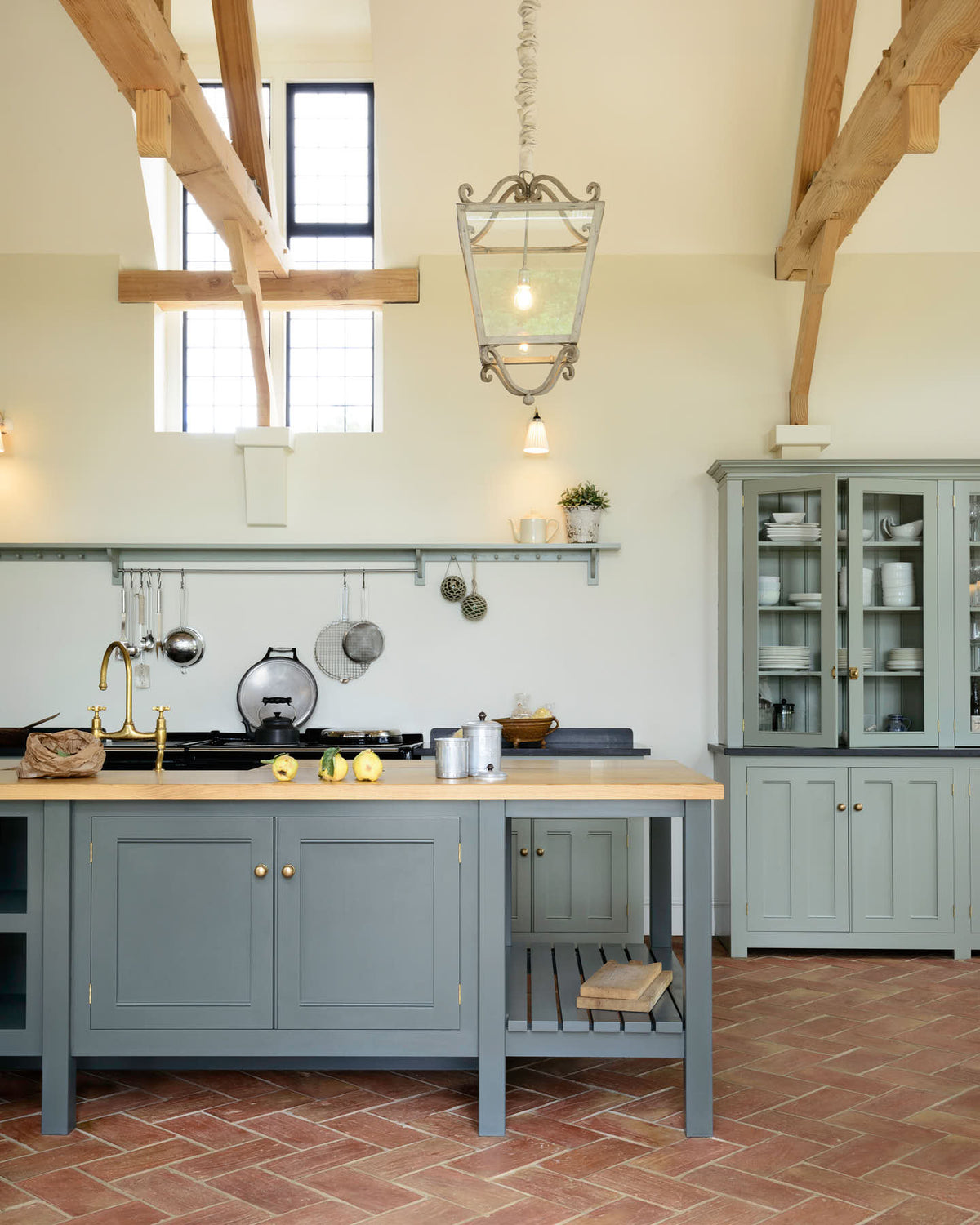 This oak-framed barn kitchen features a long cooker run with a black granite worktop, a shelf, and a kitchen island complete with a single Belfast sink and an aged brass Ionian tap atop a solid oak worktop. Adorned with framed pendant lights, artisan ceramics, books, pears, and silver kitchenware, this kitchen creates a warm and inviting space.