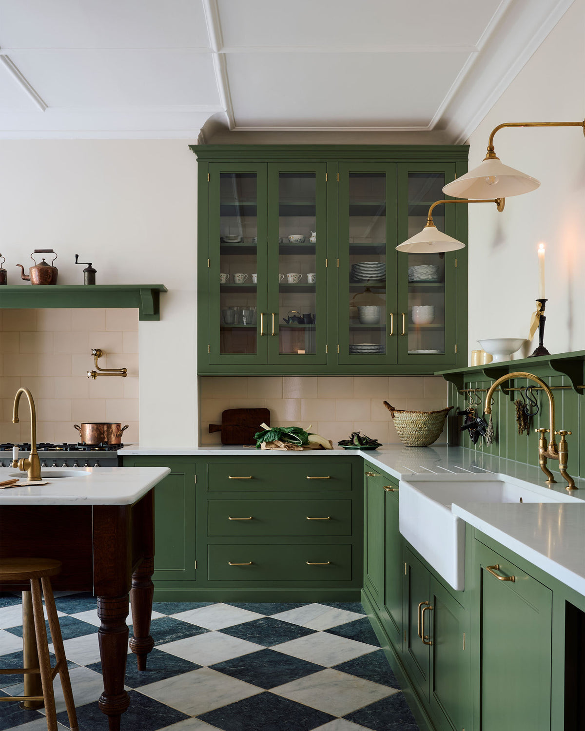A spacious and elegant Wilkes green classic English kitchen features a long L-shaped cooker and sink run with a silestone worktop, glazed wall cupboards, shelves, and an Heirloom dairy table. Adorned with classic marble chequered-board flooring, Heirloom task lights,  cream subway splash back tiles, copper pots, and luscious green vegetables, it creates a classic and stylish kitchen.