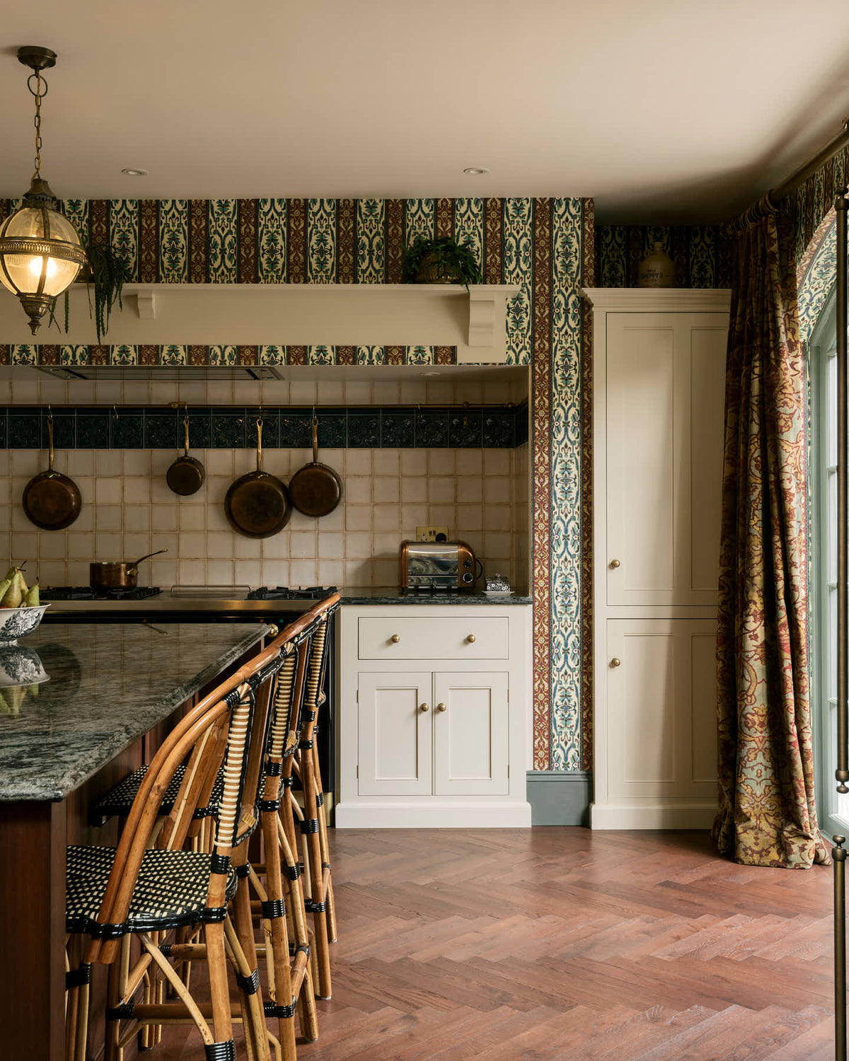 This grand and luxurious classic English kitchen features a cooker run set into the chimney breast, a large kitchen island topped with Volga blue granite, a single copper sink, an aged brass Ionian tap, and free-standing cupboards. Lavishly decorated with intricately patterned wallpaper, a William Morris pattern curtain, reclaimed splash back tiles, and hanging lantern pendant lights, it creates a royal yet comfortable and quirky space.
