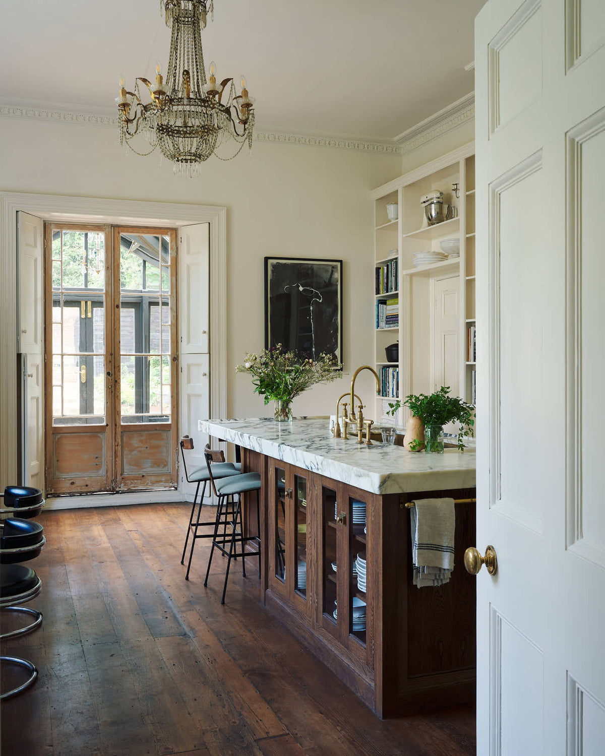 Looking through the door of a grand yet simple linen-white classic English kitchen, one sees an Arabescato marble kitchen island, a polished brass sink, and an aged brass Ionian tap. The space is simply decorated with books, glass vases filled with wildflowers from the area, and a large metal-and-crystal chandelier.