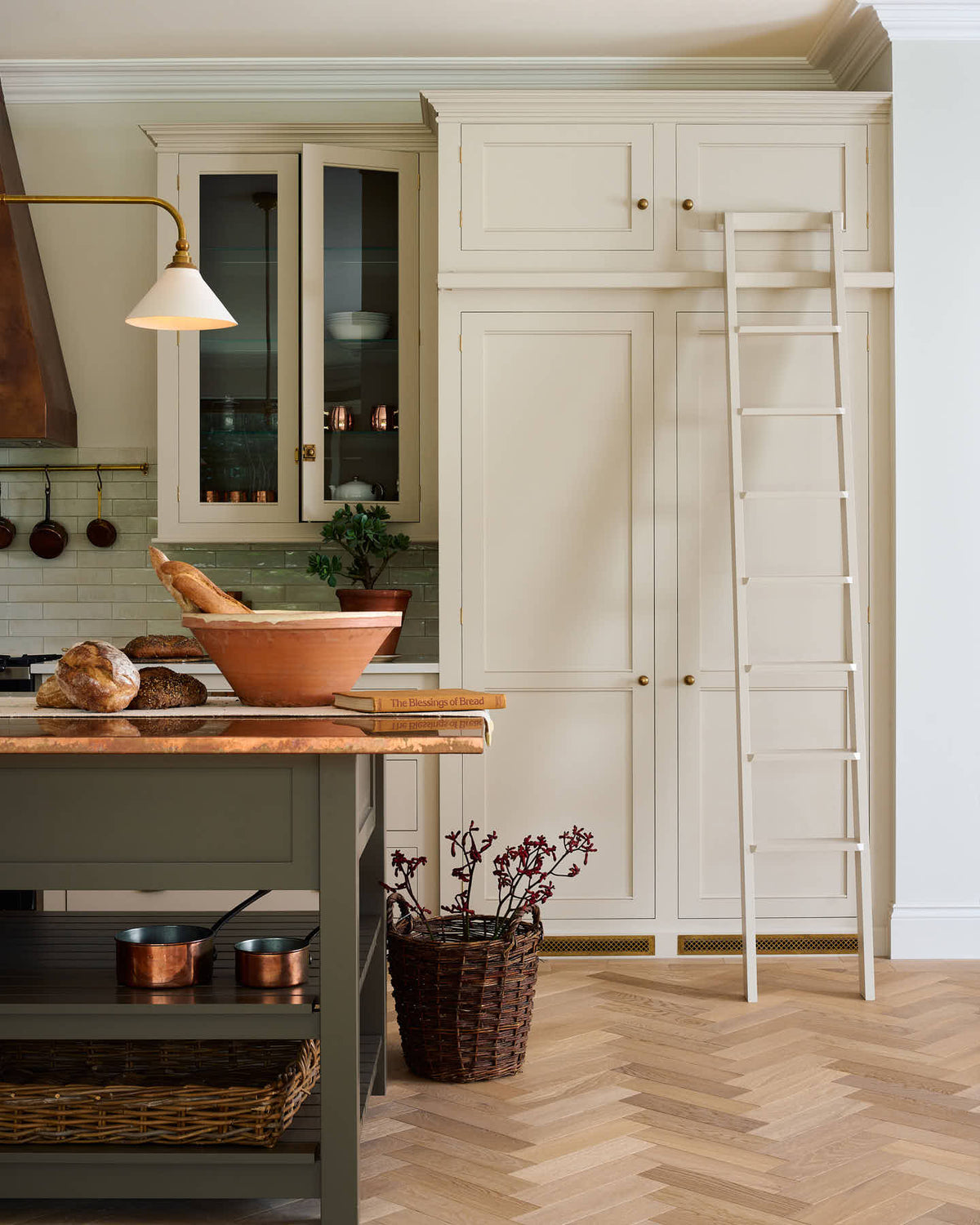 One side of a grand, earth-toned classic kitchen features a silestone worktop cooker run, glazed wall cupboards, a full-height pantry, a copper canopy, a large island with a copper worktop, and a ladder. Adorned with warm colours such as an aged brass hanging rail, deVOL Heirloom gaselier lights, textured woven baskets, vintage ceramics and loaves of bread, it makes a spectacular use of space but also down-to-earth.