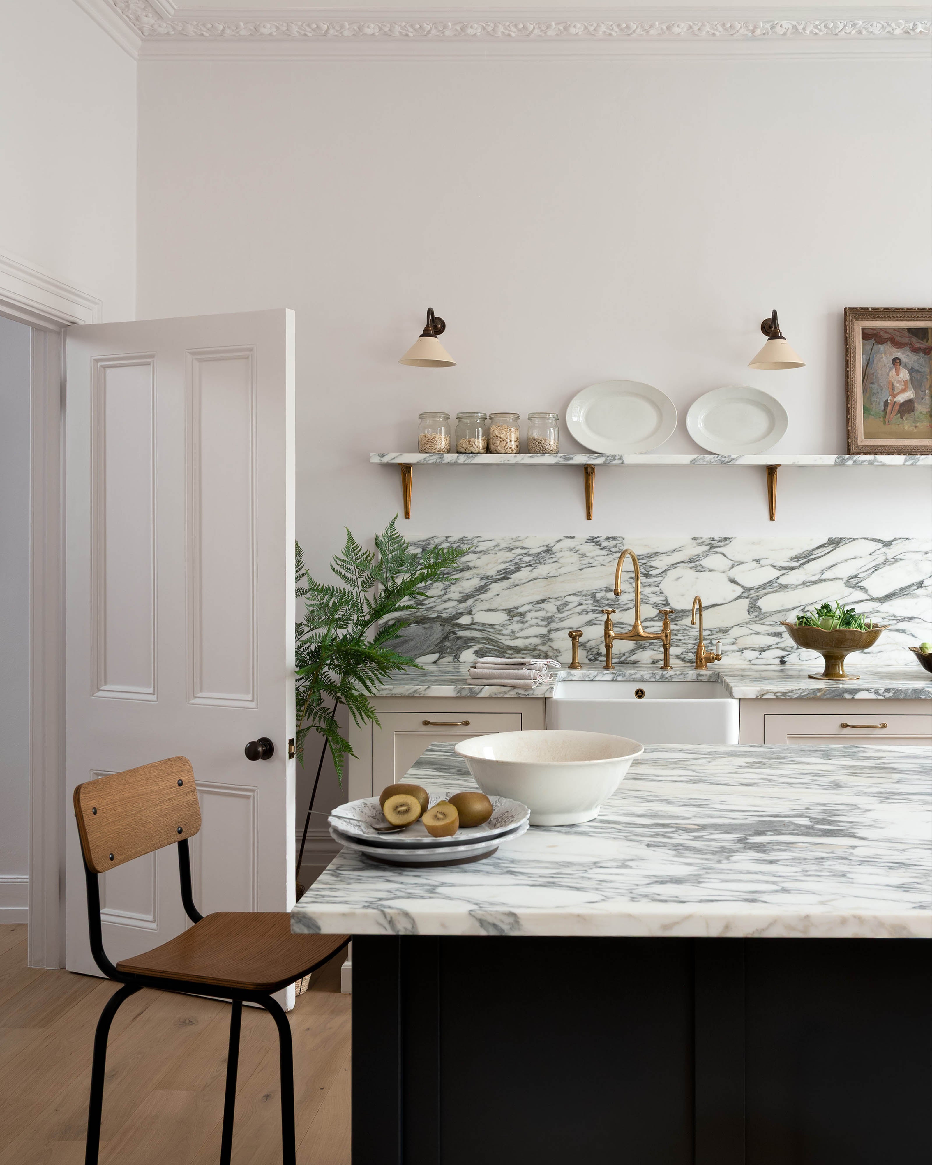 A quiet corner of a humble yet elegant off-white Classic English kitchen features a sink run with honed Arabescato marble worktop and splashback, a pantry blue kitchen island with a cooker, mini creamware wall lights, and a plumber’s stool. Adorned with artisan ceramics, brass pans, a vintage painting, fresh greens, and grains, creating an intimate social and practical kitchen space.