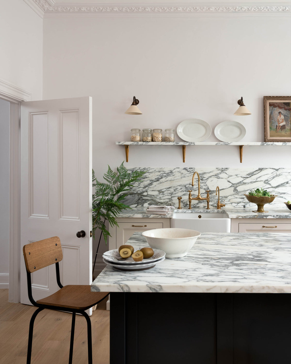 A quiet corner of a humble yet elegant off-white Classic English kitchen features a sink run with honed Arabescato marble worktop and splashback, a pantry blue kitchen island with a cooker, mini creamware wall lights, and a plumber’s stool. Adorned with artisan ceramics, brass pans, a vintage painting, fresh greens, and grains, creating an intimate social and practical kitchen space.