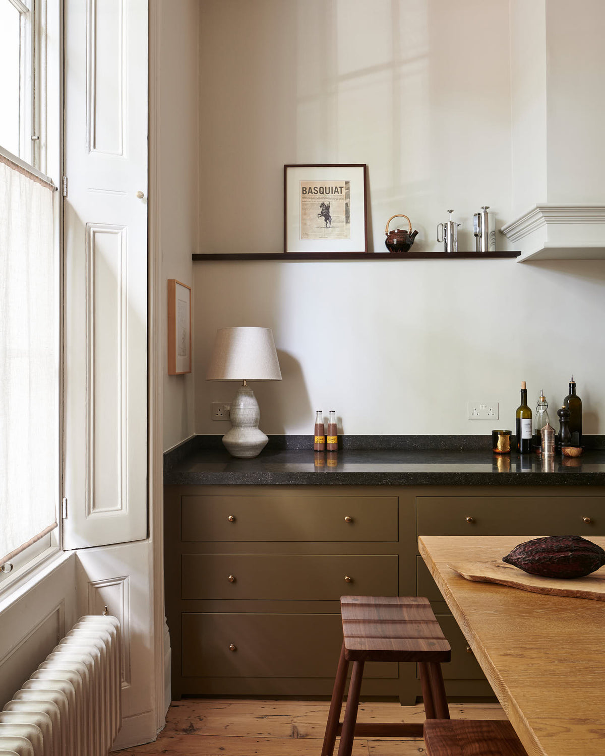 A quiet corner of an elegant olive-brown classic kitchen features a cooker run topped with Belgian blue limestone, a kitchen island with a sink and a solid oak worktop, a shaker canopy, and a built-in shelf. The tall-ceilinged room is decorated with slender bottles of wine and vinegar, a pepper grinder, a cocoa pod, and small, delicate framed illustrations.