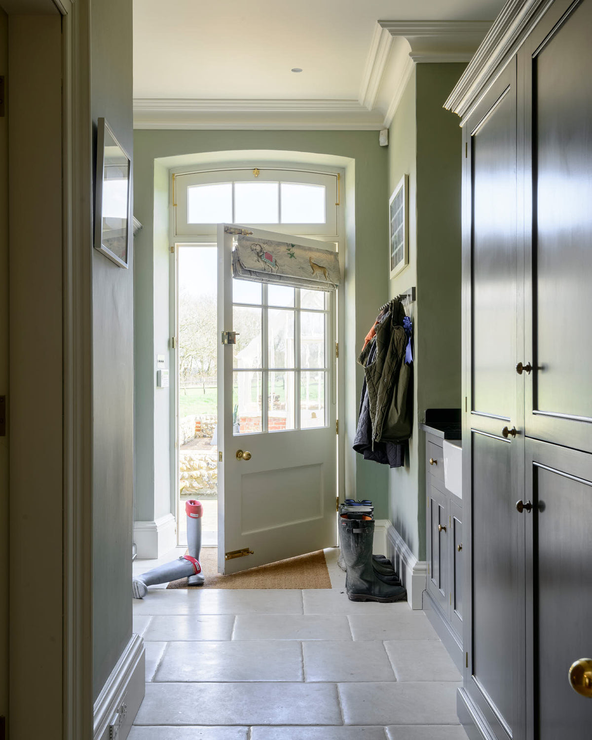 A functional muted-grey boot room features an L-shaped bench, shelves, a peg rail, a small sink run with a single Belfast Ionian sink, a black granite worktop, and full-height storage cupboards. Beautifully decorated with thick coats and muddy boots, this space leads into the back garden, creating a practical and family-oriented area.