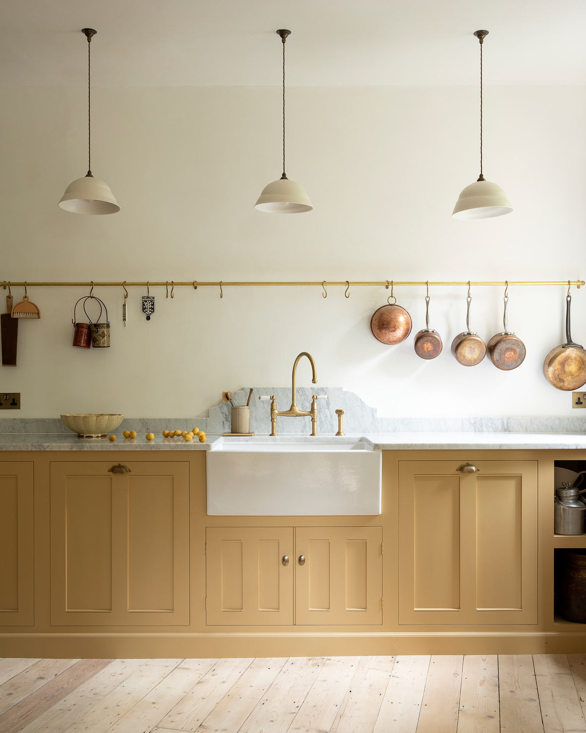 This charming blush yellow, classic English kitchen features a Carrara marble countertop, an ornate backsplash, and an aged brass tap. Decorated with copper pots, vintage ceramics, and small wooden cleaning supplies, it creates an elegant and tidy atmosphere.