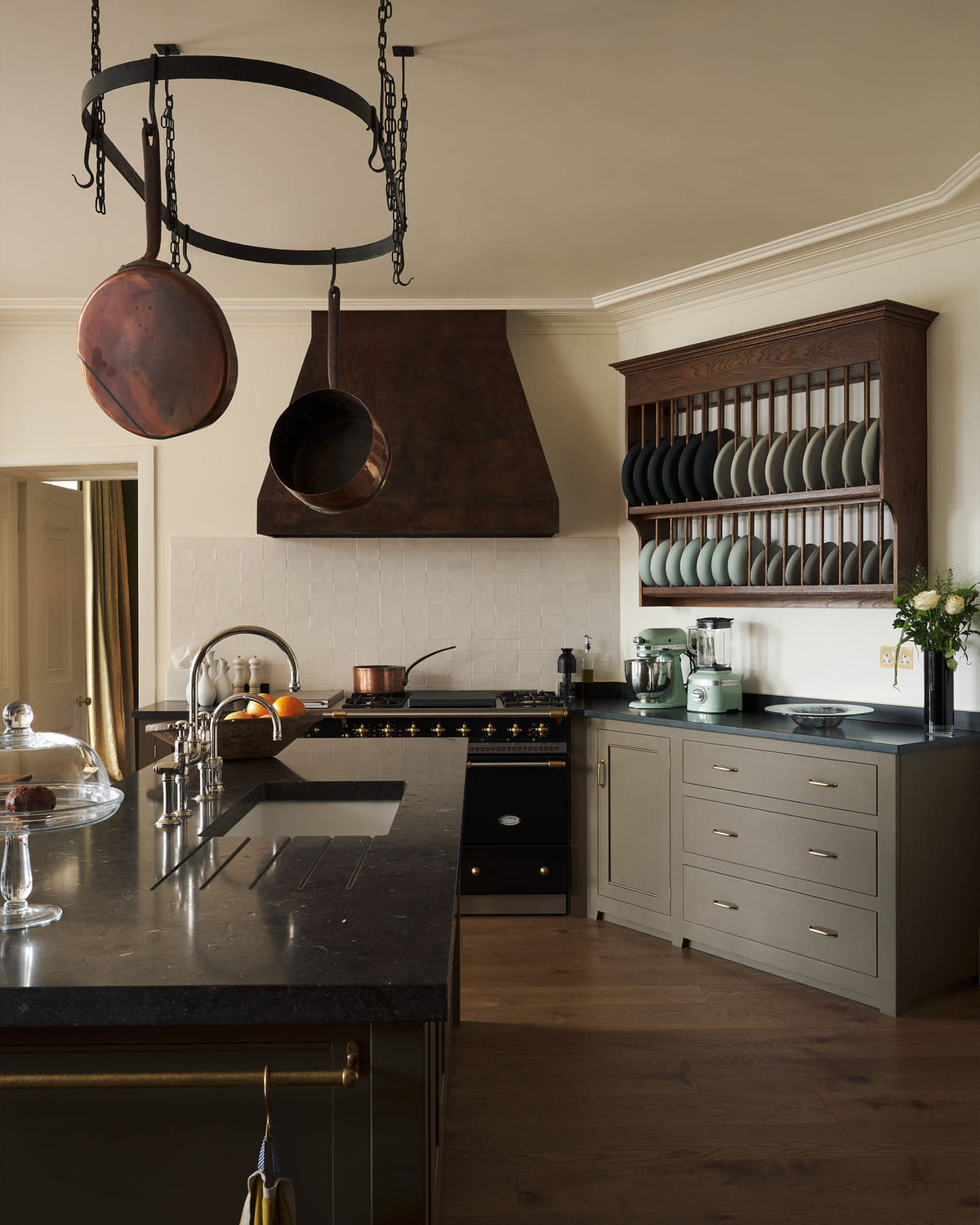 A spacious olive-green classic kitchen features a large island with a Belgian blue limestone worktop, a single sink, and an antique silver Ionian tap. Above, a deVOL pot hanger takes centre stage. The classic cooker run showcases a large black gas stove, a substantial canopy, and an heirloom plate rack displaying colour-coordinated muted greens and dark blue ceramic plates.