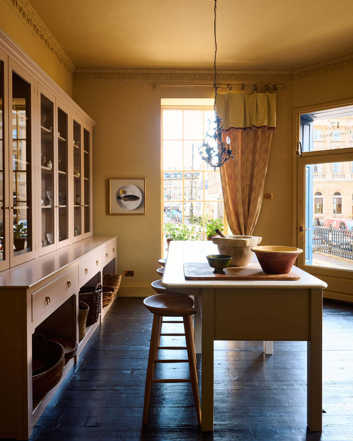 A warm yellow room with a large Princelet pink dresser showcasing glazed cupboards filled with delicate ceramics, papier-mâché mushrooms, and wooden baskets along the bottom, giving the space a relaxed, living-room feel. The space includes a wide prep table with an open nook for deVOL bum stools, there’s also a vintage armchair sofa, large ceramic bowls, a metal vines-and-flowers chandelier, and a golden-framed painting of a fish and a lemon.