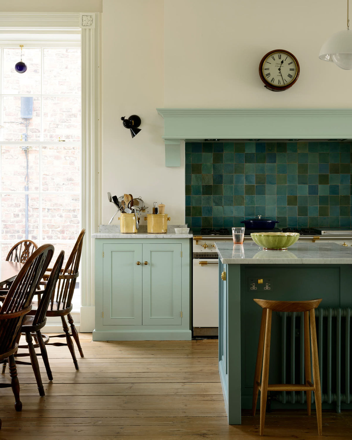 A corner of a humble and calming turquoise, classic English kitchen featuring a cooker and sink run with a Carrara marble worktop, a suspended false chimney breast, a glazed wall cupboard, and a kitchen island with Carrara marble on one side and solid oak on the other. Adorned with symmetrical hanging lights, butter-yellow ceramics, deVOL bum stools and ocean porcelain tiles, it creates a warm and simple home kitchen.