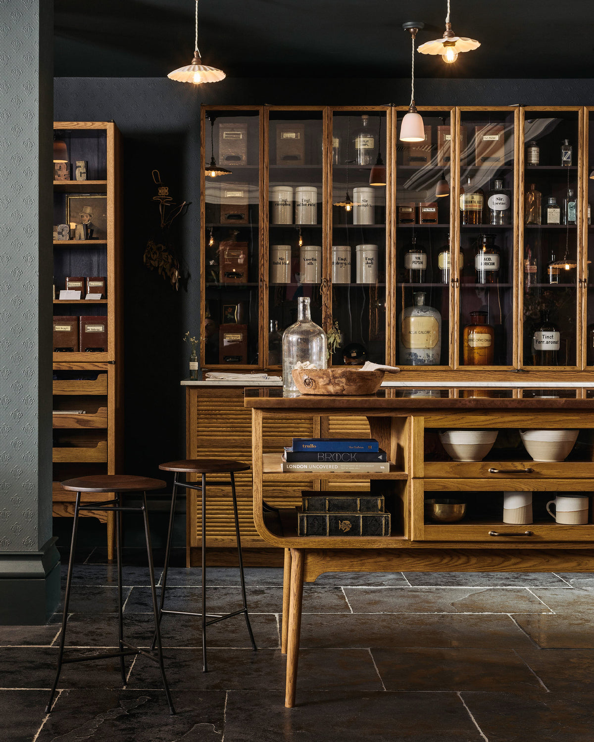 A dark room with charcoal-coloured walls and ceiling and a brushed charcoal limestone floor, contrasting with the wooden haberdasher's kitchen furniture. In the foreground, there are two creedy stools and a prep table with turned oak legs, an open curved end, glazed drawers full of rustic pottery and a mottled copper worktop. Behind is a tall bank of glazed countertop cupboards full of antique chemists' jars and tins, each with a handwritten label.