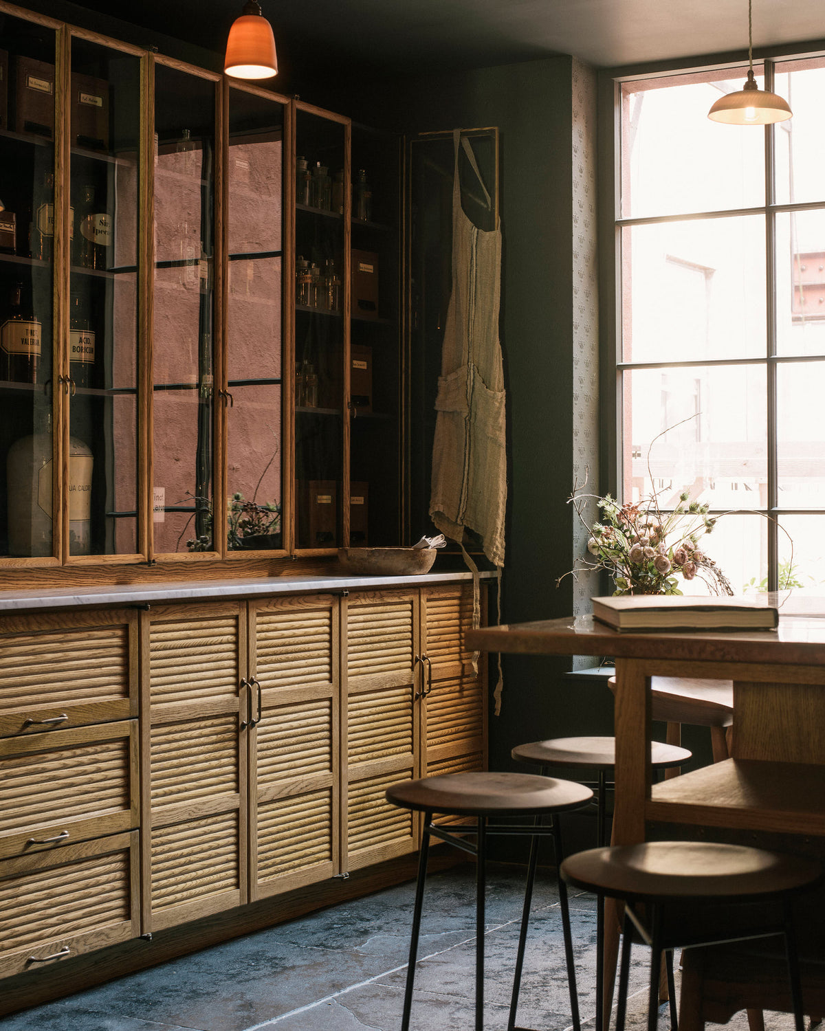 An oblique view of a bank of Haberdasher's tall glazed countertop cupboards with skinny oak frames above a run of tambour-style slatted base cabinets. The cupboards are made from oak with an amber-coloured and slightly aged oil finish. Light pours across the front of the cupboards from a tall window to the right of the run.