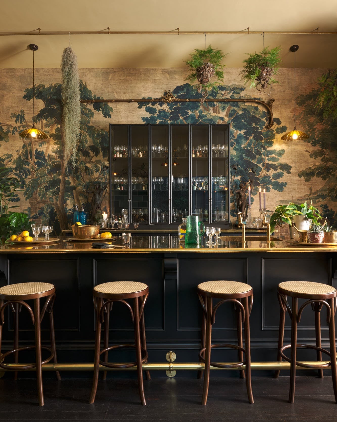 A chic and glamorous Bobbin Black Haberdasher’s bar featuring a kitchen island with a polished brass worktop, a single polished brass sink, aged brass Mayan taps, a brass foot rail, glazed base cabinet with internal drawers, tall countertop cupboards, and woven top bentwood café stools. Set against an illustrated foliage wallpaper and adorned with hanging plants, wine glasses, and vintage decanters, it creates a beautiful and intimate personal bar.