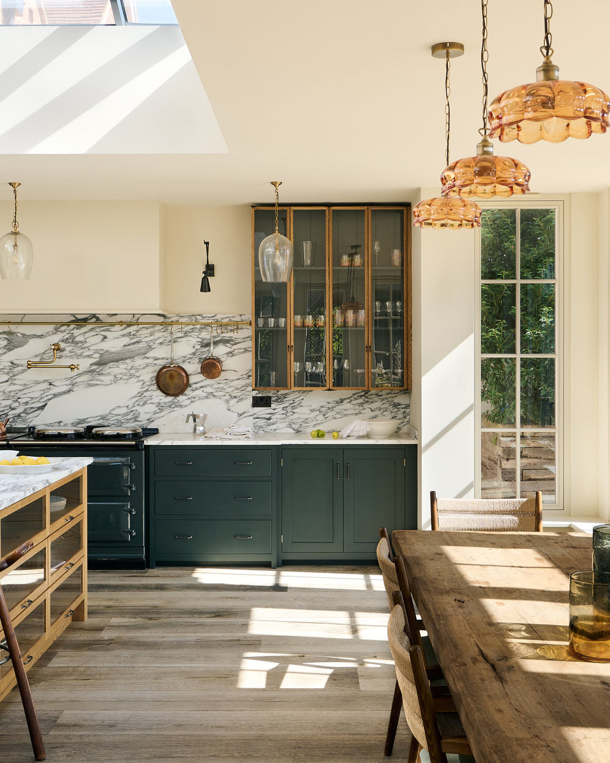 A quiet corner of an elegant and spacious Haberdasher’s kitchen features a large cooker run, a kitchen island with an Arabescato marble worktop, a splashback, and glazed cupboards. Adorned with vintage furniture, artisan ceramics, glass pendant lights, copper pans, and a bowl of lemons, this kitchen creates a classic social space with functional storage.