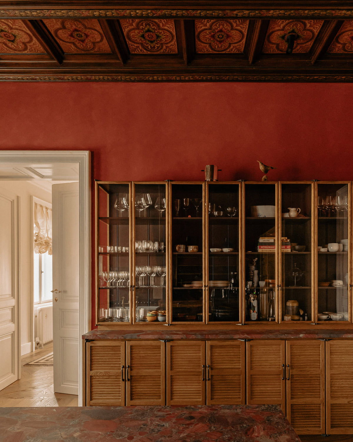On the other side of a warm and historical Haberdasher’s kitchen features a pantry run featuring glazed countertop cupboards, a kitchen island, and a red marble worktop. Beautifully decorated with floral ceiling tiles, books, clear drinking glasses, and artisan ceramics, this kitchen creates a grand yet comfortable space.