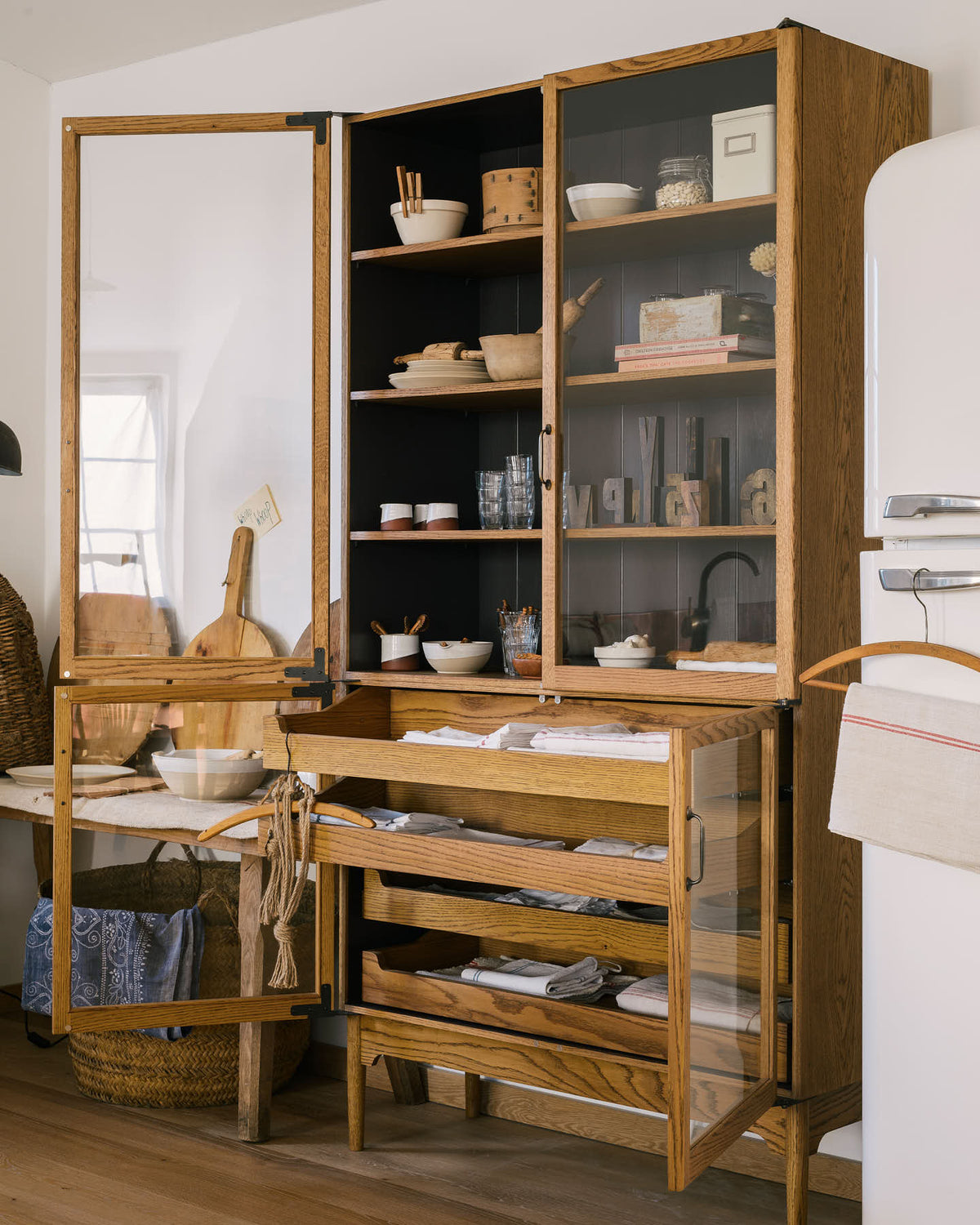 A closer look at a warm and spacious Haberdasher’s kitchen featuring a freestanding pantry with glazed doors. Adorned with vintage furniture and items, artisan ceramics, wicker baskets, tea towels, large wooden tapas boards, and wooden letterpress, this kitchen creates a contemporary yet nostalgic space.