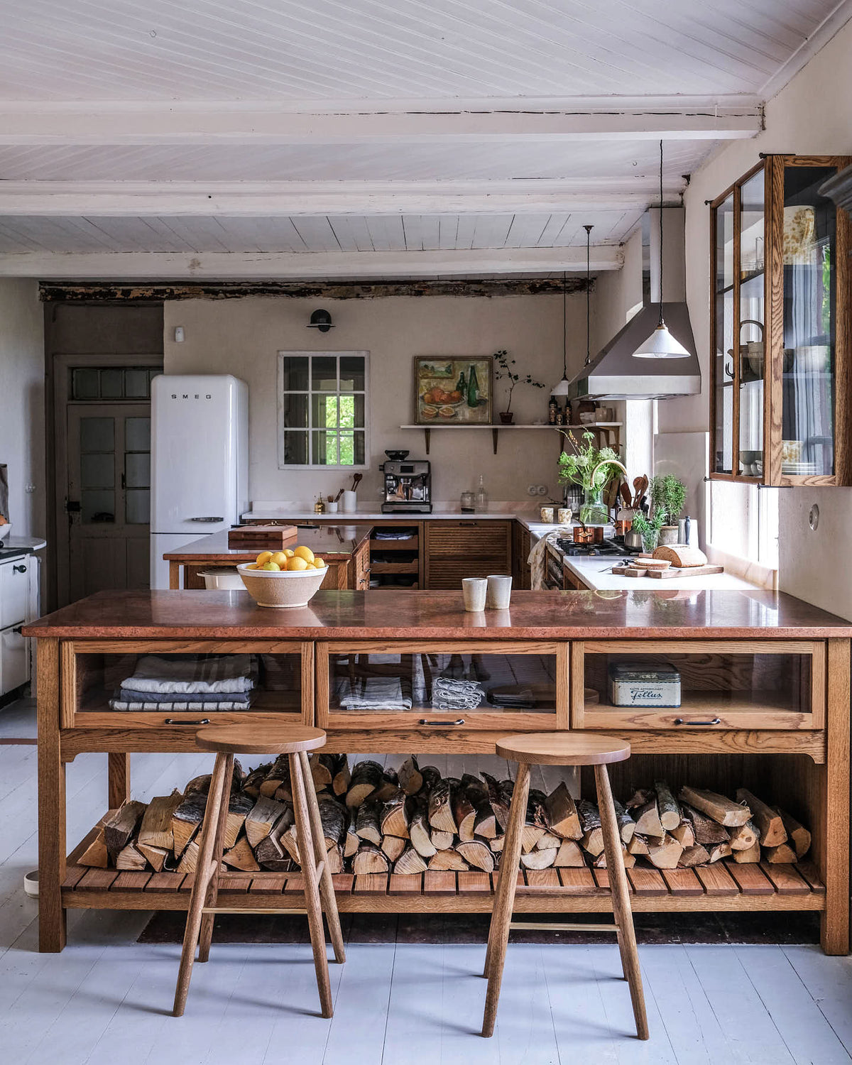 A warm and inviting Haberdasher’s kitchen features a sink and cooker run with a Carrara marble worktop, a double-fluted marble sink, an aged brass Ionian tap, prep tables with a copper worktop, glazed wall cupboards, and a shelf. Beautifully decorated with vintage furniture and items, artisan ceramics, firewood, Helen stools, and a bowl of lemons, this kitchen creates an airy and cosy Scandinavian small fishing town hideaway.