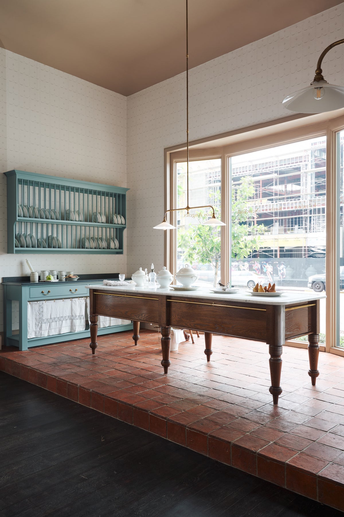 An oblique view through a door way of a beautiful and functional Trinity blue Heirloom kitchen features open cabinets, plate racks, and a showpiece Dairy table. Lovingly adorned with bone white artisan ceramics, selected antiques, lace curtains, Gaselier lights, and earthy red terracotta square floor tiles that recreate Victorian kitchen furniture that is made to last a lifetime.