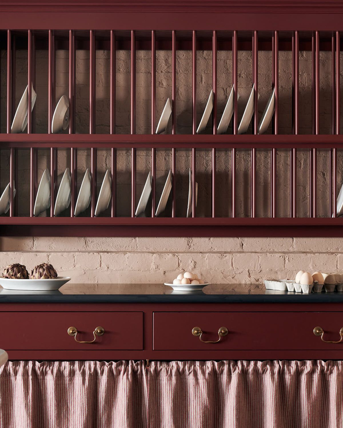 A closer look at the refectory-red Heirloom kitchen’s sink cabinet reveals  café curtain rails, a red and white striped curtain, and a plate rack. Beautifully decorated with artisan ceramics and plates of fresh eggs, this kitchen offers a modern twist on vintage furniture, creating a classic range designed to last.