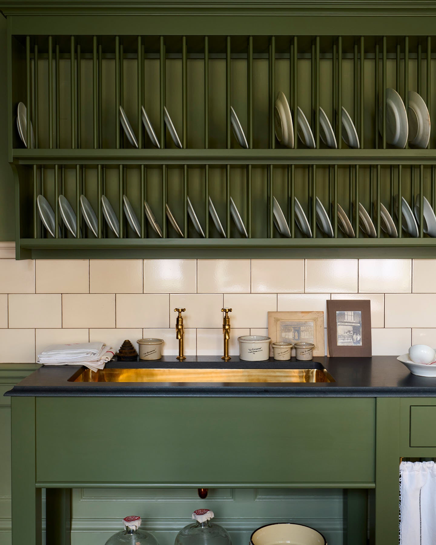 An closer look at the quaint and stylish Wilkes green Heirloom kitchen features a sink cabinet with a café rail, white lace curtains, a polished brass sink, aged brass Mayan taps, and a large plate rack. Adorned with vintage photos, a cream bucket, simple ceramics, cream subway splash back tiles, and a golden-framed sliced green pumpkin, this kitchen creates a luxurious dining and social space.