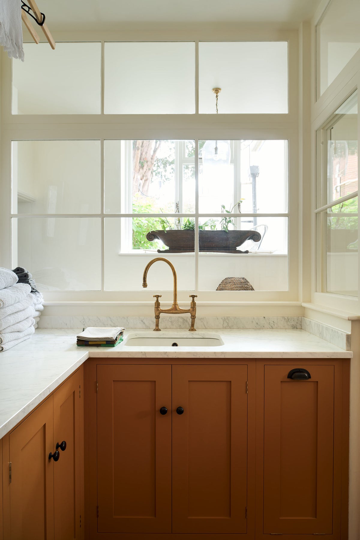 A closer look at a scullery yellow laundry room reveals an L-shaped sink with a white marble countertop and a stunning aged brass Ionian faucet. Neatly stacked white towels are arranged in layers, and the view through the window lends the space a beautiful sense of calm.
