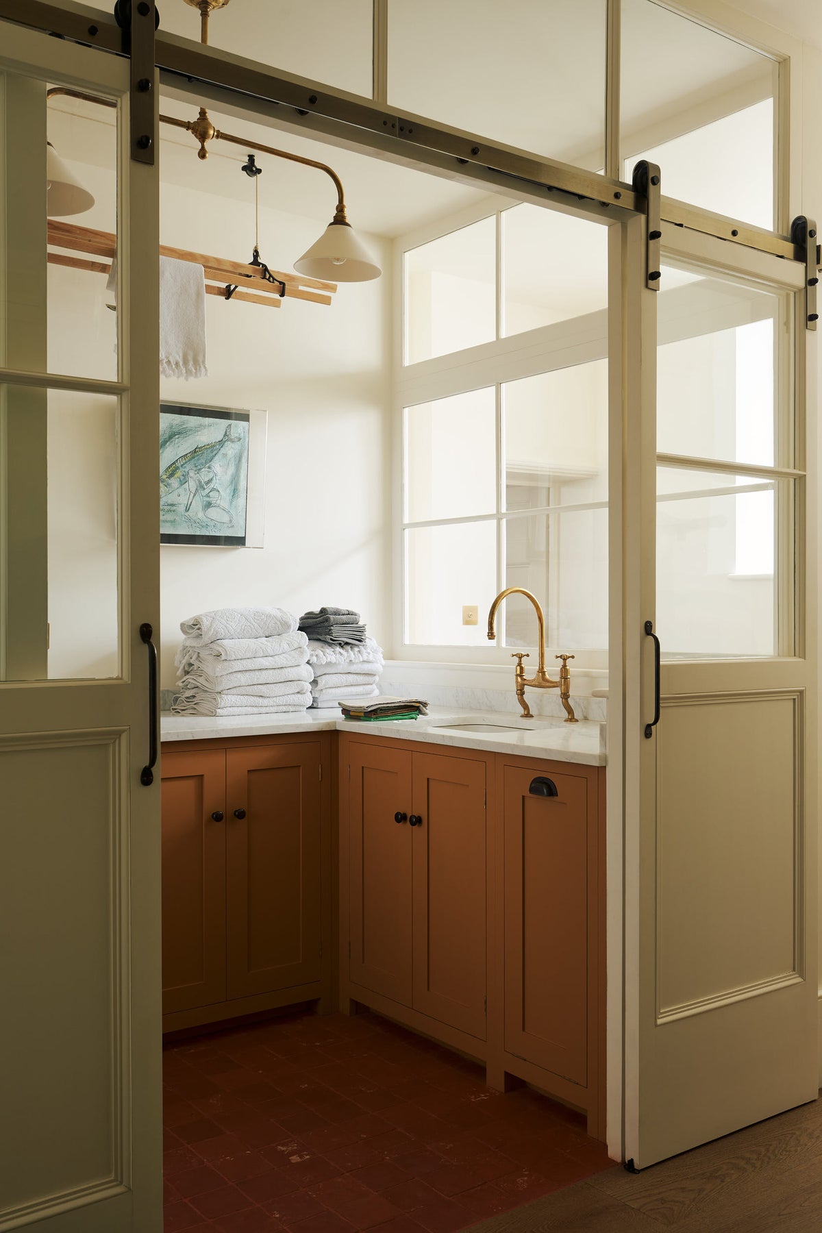 An oblique view of a cream sliding door that leads to a scullery yellow laundry room featuring an L-shaped sink with a white marble countertop and a stunning aged brass Ionian faucet. Neatly stacked white towels are arranged in layers, and two framed artworks adorn the walls. In the centre of the ceiling hangs a deVOL Gaselier light, while a deVOL Laundry Maid, with a towel draped over it, is positioned behind.