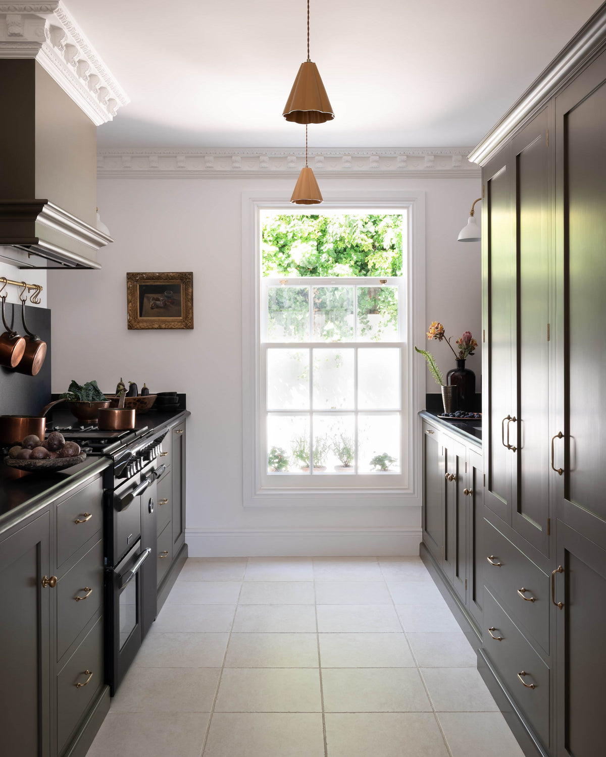 Looking through a simple archway is a murky-green galley shaker kitchen, featuring a cooker run with a black granite worktop and splashback, an aged brass hanging hook, a shaker canopy, a sink run, and pantry cupboards. Beautifully curated with two copper pendant lights, a vintage gold-framed painting, artisan ceramics, fresh produce, houseplants, and a wide open window, it creates a chic, understated, and practical galley kitchen.