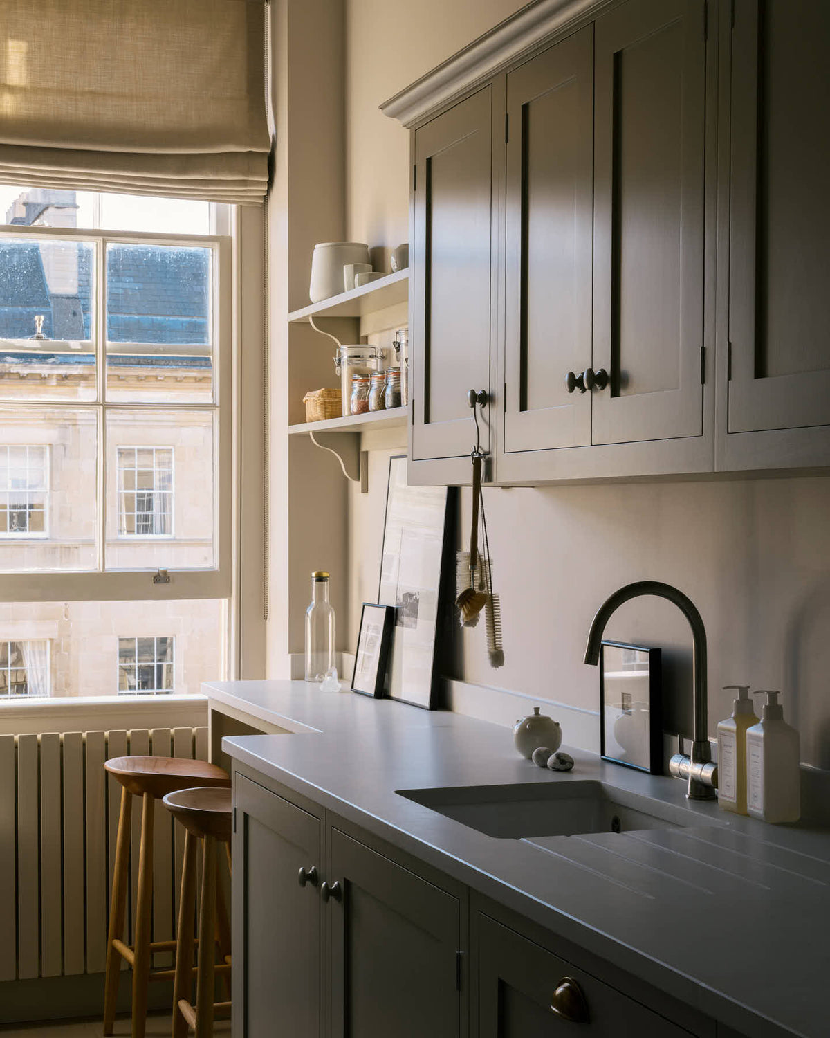 An angled view of a stylish, mushroom-coloured Shaker galley kitchen features a cooker and sink run with silestone worktop, wall cupboards and a cosy breakfast nook. Beautifully decorated with simple, everyday kitchen items such as dried ingredients, muted ceramics, framed illustrations, deVOL Bum Chairs, a healing crystal, and an amazing view of Bath’s famous Georgian apartments, this calming and functional kitchen space blends character with peace.