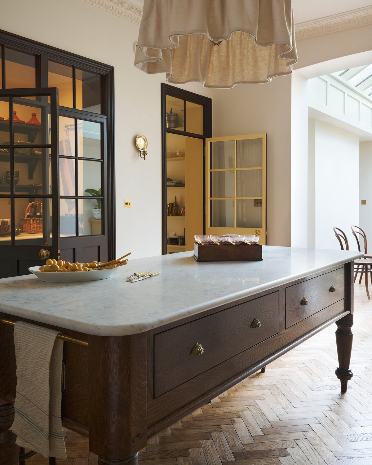 A closer look at the large Dairy Table in the soft Princelet pink Shaker kitchen, highlighted by a grand chandelier with a loose fabric shade and glazed doors leading to a spacious pantry.