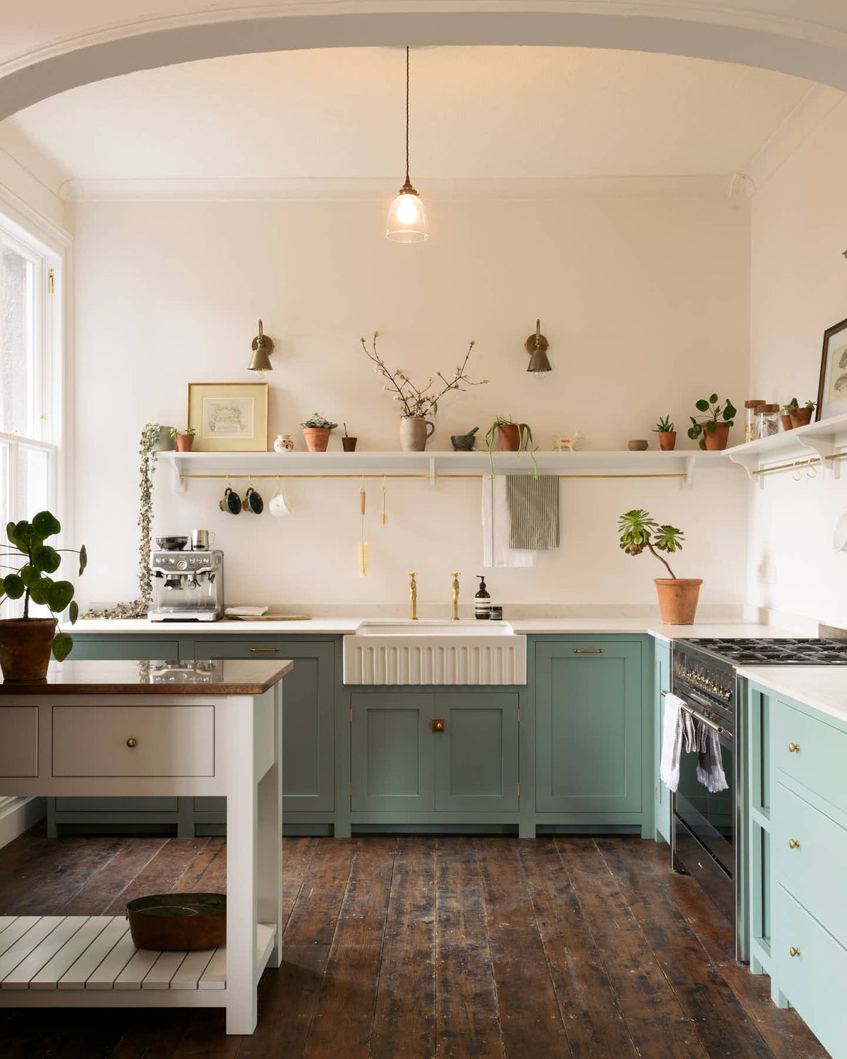 A close look at a freestanding prep table in linen white with a copper worktop in the Trinity Blue shaker kitchen with an L-shaped cooker and sink run. The kitchen features a countertop cupboard with a Carrara marble top, a long shelf with brass rails for hanging cups, cleaning tools, and towels.