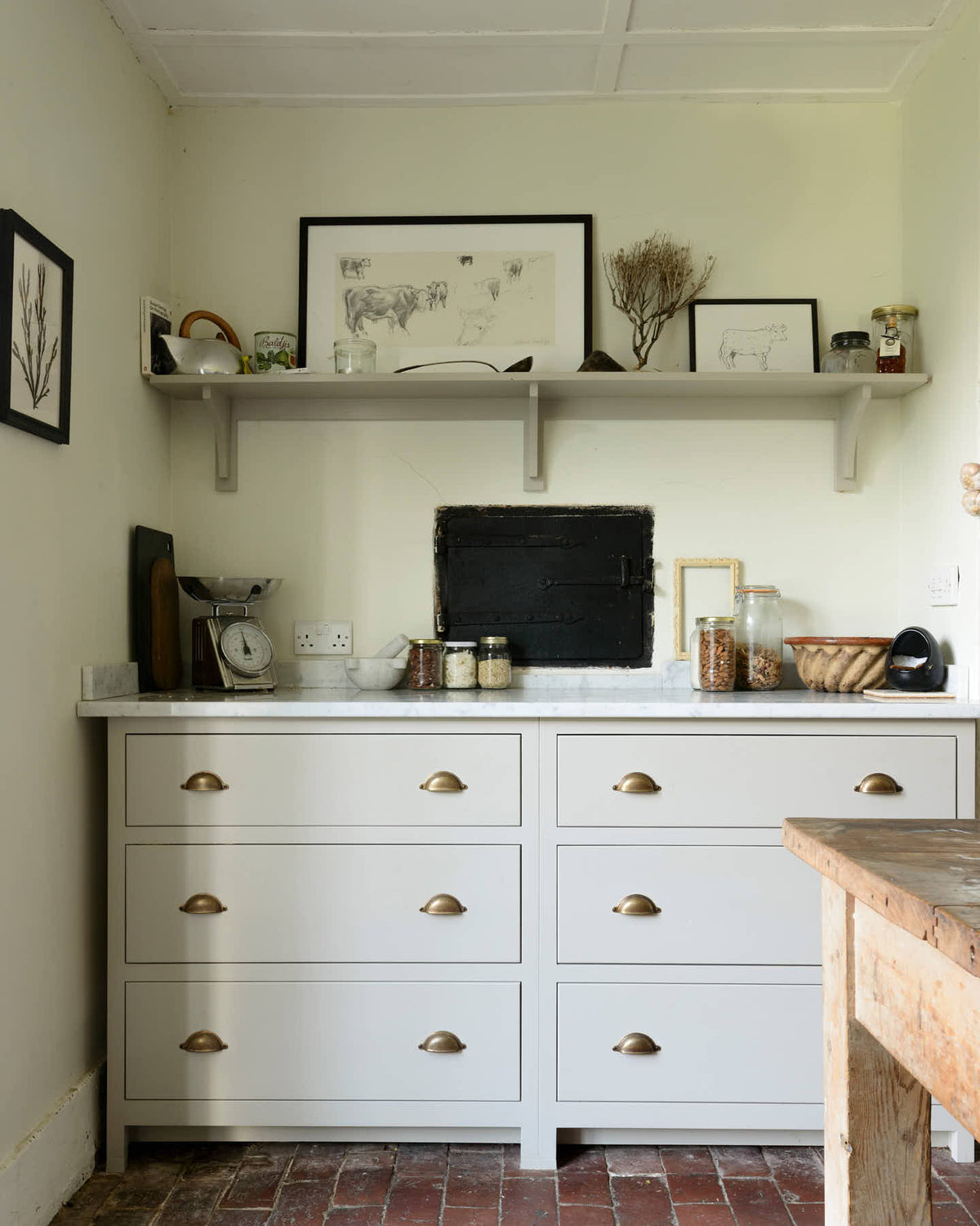 A quiet corner of a small and quaint mushroom-coloured Shaker kitchen features a sink run with Carrara marble worktop. Beautifully decorated with vintage furniture such as a large solid oak table, a silver scale, and assorted bric-a-brac, it creates a lovely social space full of character.